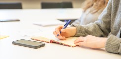 Studierende sitzen mit Stiften in der Hand an Tischen und blicken nach vorn