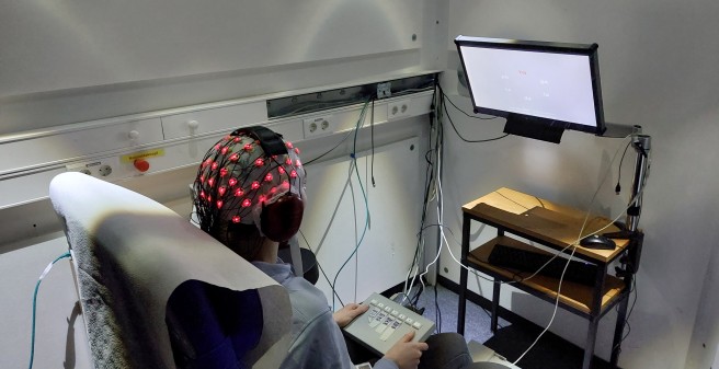 Mensch sitzt für EEG-Messung mit Elektroden auf dem Kopf in Untersuchungsraum und guckt auf Bildschirm
