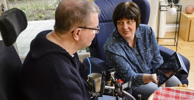 Tomas Gerlach im Rollstuhl und seine Frau unterhalten sich, sie zeigt ihm etwas auf dem Monitor