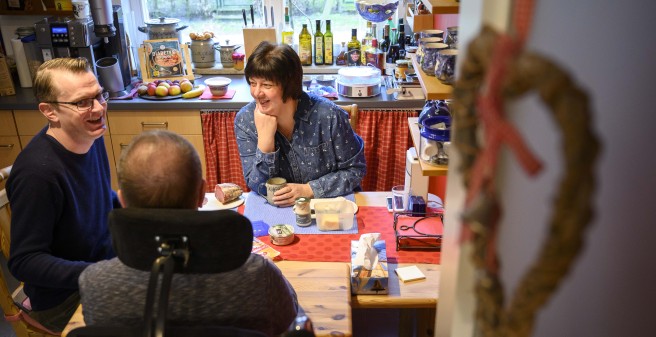 Tomas Gerlach im Rollstuhl mit Ehefrau und Betreuer lachend am Frühstückstisch