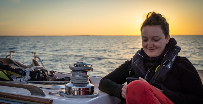 Sonnenuntergang über der See, eine Frau lehnt an der Reeling und schaut versonnen lächelnd auf ihr Handy