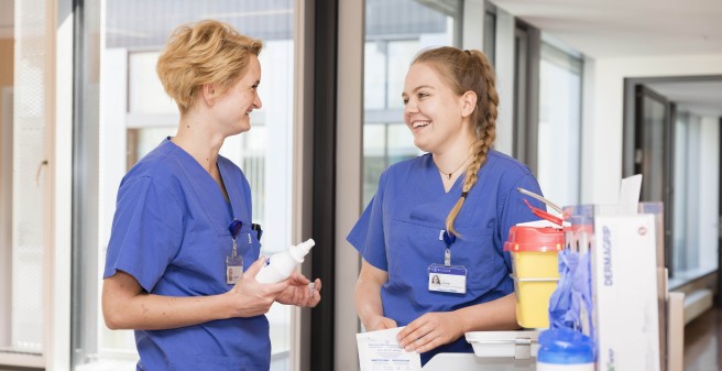 Eine junge Pflegerin im Gespräch mit ihrer Kollegin, beide lächeln sich freundlich an