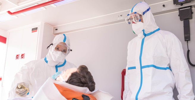 Stefanie_Beck und ihr Kollege, Notfallsanitäter Sebastian Götsch, komplett in Maske und weißen Anzug sind mit einem Patienten im Rettungswagen.