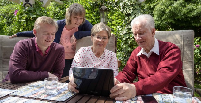 Peter Seifert und seine Familie sitzen glücklich auf der überdachten Terasse und blicken in einen Monitor. Ein sonniger Tag.