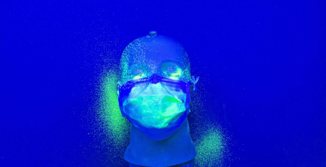 Auf einem tiefblauen Untergrund liegt ein Kunststoff-Kopf mit einer Mund-Nasenschutz-Maske. Auf der Maske, sowie links und rechts auf der Unterlage sind hellgrüne Punkte verstreut