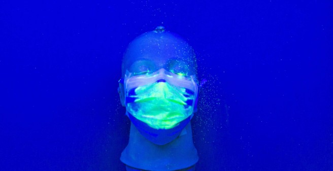 Auf einem tiefblauen Untergrund liegt ein Kunststoff-Kopf mit einer Mund-Nasenschutz-Maske. Auf der Maske sind hellgrüne Punkte
