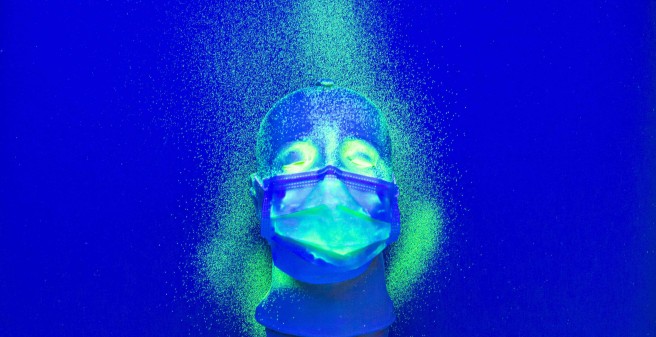 Auf einem tiefblauen Untergrund liegt ein Kunststoff-Kopf mit einer Mund-Nasenschutz-Maske. Auf der Maske, dem oberen Teil des Gesichts und Keilförmig links und oben sind hellgrüne Punkte