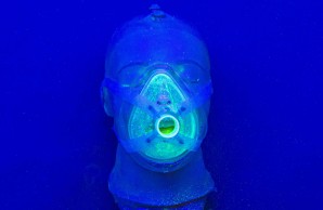 Ein Kunstoffkopf liegt auf einem tiefblauen Untergrund. er trägt eine spezielle Atemschutzmaske aus Kunststoff. Hellgrüne Tröpfchen befinden sich innerhalb der Maske, wenige im Hals/Brustbereich