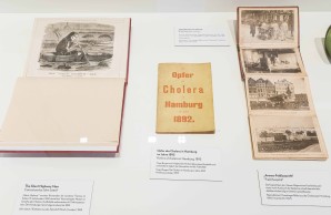 Exponate der Ausstellung zur Colera in Hamburg von 1892. Verschiedene Fotos und Zeichnungen werden in einem Schaukasten gezeigt.