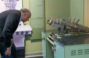 Prof Osten blickt in ein historisches Elektronenmikroskop. An der Wand hinter ihm eine technische Zeichnung und Erläuterungen