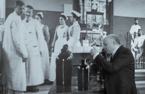 EIn s/w Bild. Prof Osten vor einem historischen Foto eines Krankensaals. Prof Osten betrachtet die vor dem großen Bild ausgestellten Flaschen