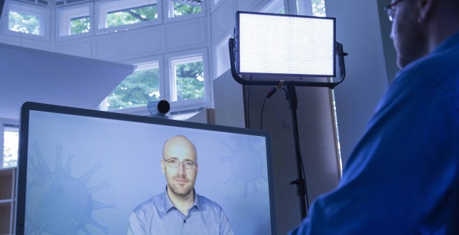 David Rickert sitzt vor einem Monitor auf dem er selbst zu sehen ist. Auf dem Monitor eine Webcam, dahinter ein Scheinwerfer