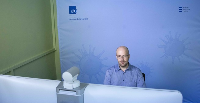 David Rickert sitzt vor einem Monitor in einem ausgeleuchteten Aufnahme-Studio. Hinter ihm eine Leinwand mit stilisierten Viren und dem UKE Logo.
