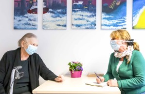 Frau Gerber und Patientin Frau Trieb, beide mit Atemschutzmasken, sitzen am Tisch, einander aufmersam zugewandt