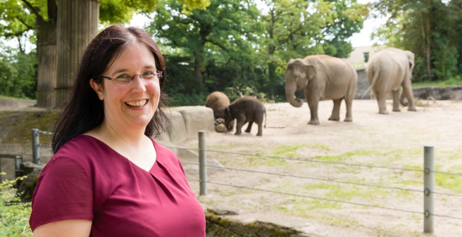 Prof. Karin Oechsle lächelt in die Kamera, hinter ihr Elefanten im Gehege