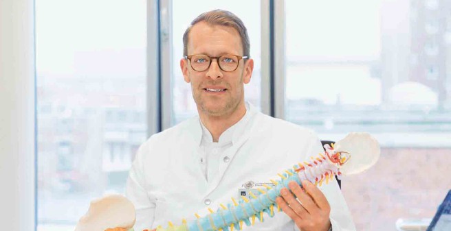 Dr. Sven Eickert hält eine Wirbelsäule aus Kunststoff mit Becken und Kopfschale in den Händen, einige Bereich sind farbliche gekennzeichnet