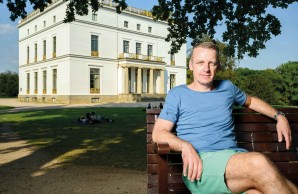 Prof. Stefan Kluge sitzt lässig auf einer Bank im Park, hinter ihm die goldverzierte, weiße Villa des ehemaligen Senator Marin Johann Jenisch