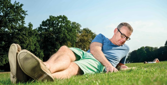 Prof. Kluge liegt lässig auf dem Rasen, er liest ein Buch, im Hintergrund seine Frau