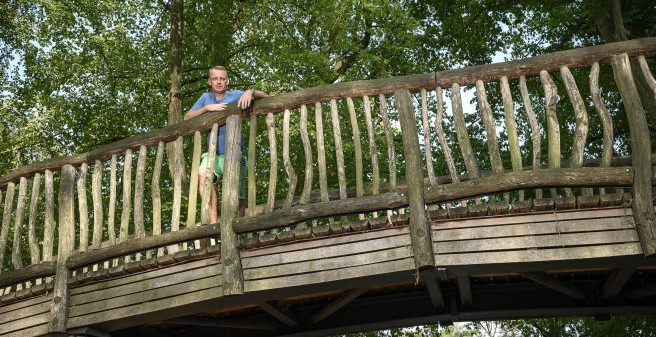 Prof. Stefan Kluge blickt über das Geländer einer urigen Holzbrücke in die Kamera. Das Geländer der Brücke bilden unregelmaäßig gewachsene schmale Stämme