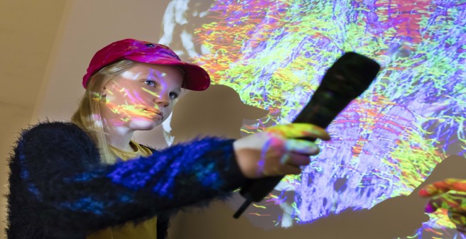 Kinderreporterin Jonna mit Mikrofon vor der Projektion einer bunt gefärbten Aufnahme des Gehirns