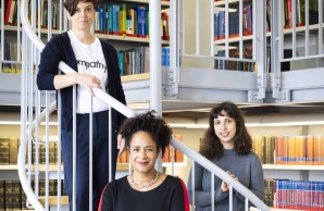 (v.l.): Dr. Christine Dahlke, Prof. Dr. Marylyn Addo, im Vordergrund, und Dr. Anahita Fathi stehen au feiner Wendeltreppe in einer Bibliothek und blicken lächelnd in die Kamera