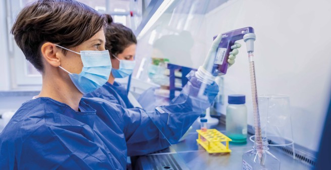 Dr. Christine Dahlke (vorn) und  Marie Weskamm in blauen Schutzkitteln, Handschuhen und Alltagsmasken pipettieren an ihrem Labor-Arbeitsplatz