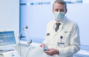 Prof. Kluge steht an einem leeren Intensivbett, einen Tubus in der Hand. Er trägt einen weißen Kittel und Alltagsmaske, Erklärend blickt er ernsthaft in die Kamera
