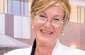 Prof. Dr. Ulrike Ravens-Sieberer lächelt mit schräg gelegtem Kopf in die Kamera, im Hintergrund ein Foto des UKE EIngangsbereichts