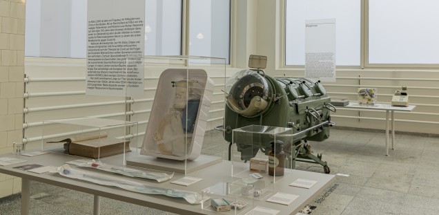  Eine Eiserne Lunge aus den 1950er Jahren zwischen Ausstellungstischen, im Vordergrund ein modernes ECMO-KIt aus der Intensivmedizin